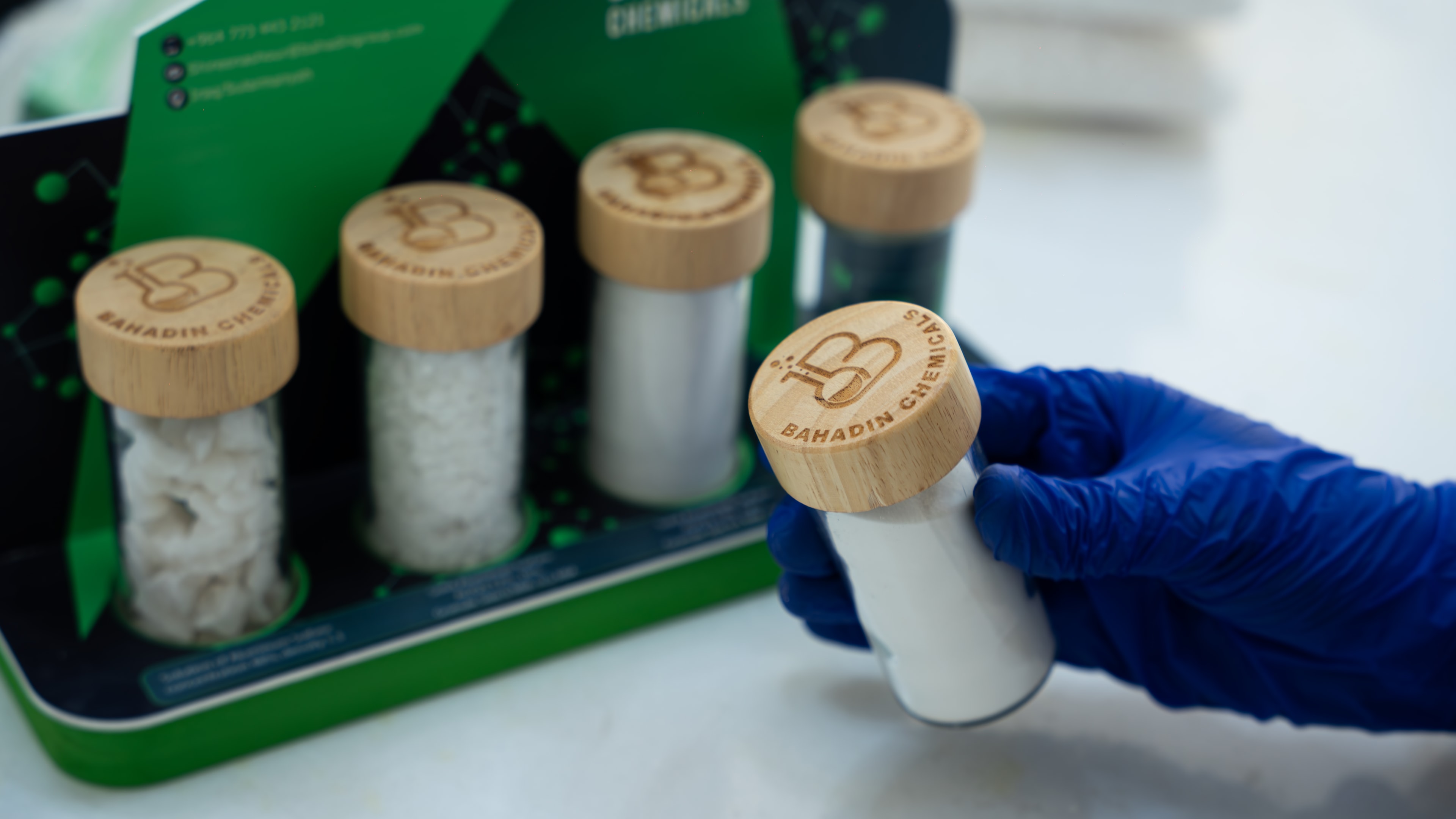 A gloved hand lifting a jar of aluminium sulfate, its lid branded Bahadin Chemicals