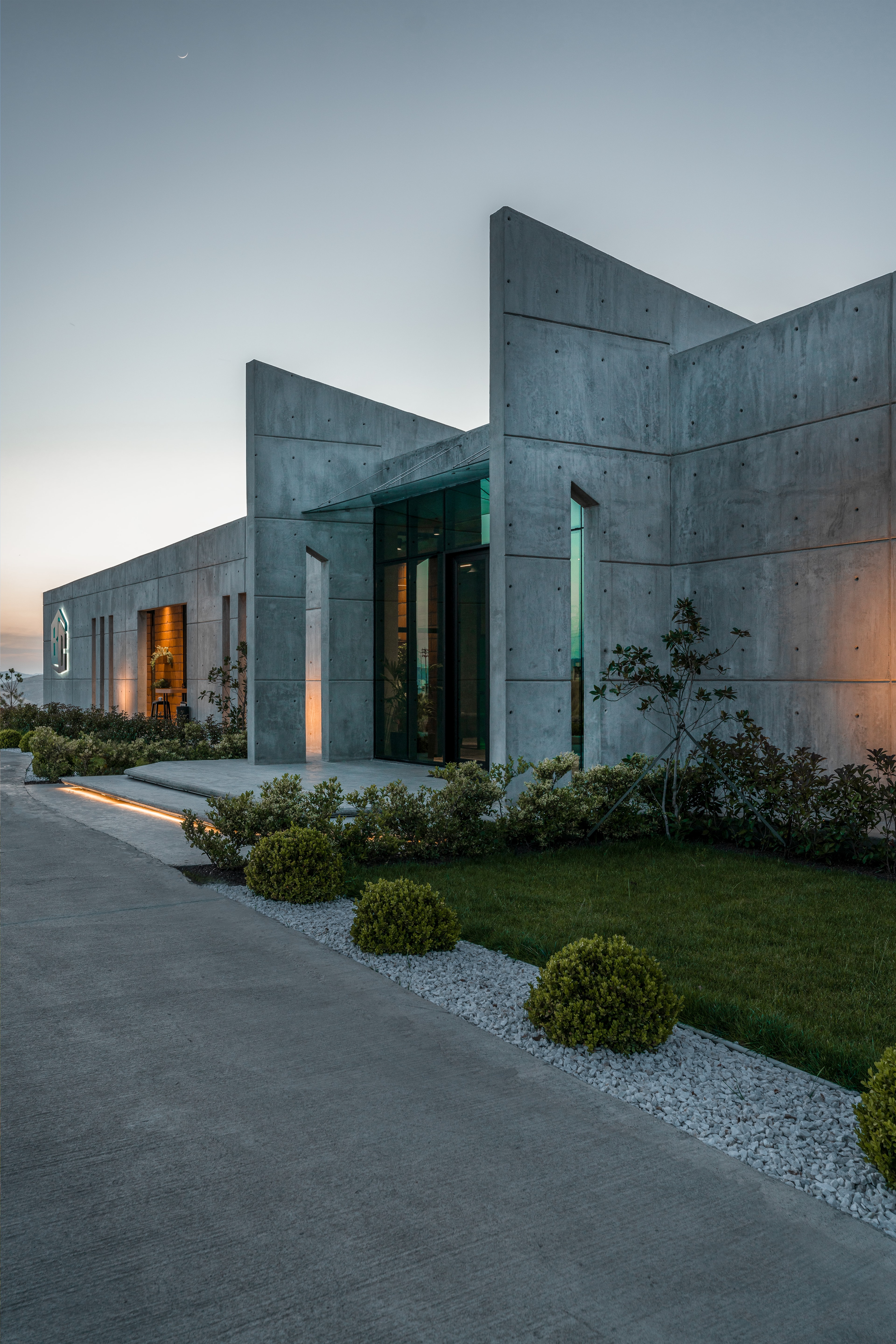 The Bahadin Concrete head office at dusk: angled concrete walls above a lit path and gardens